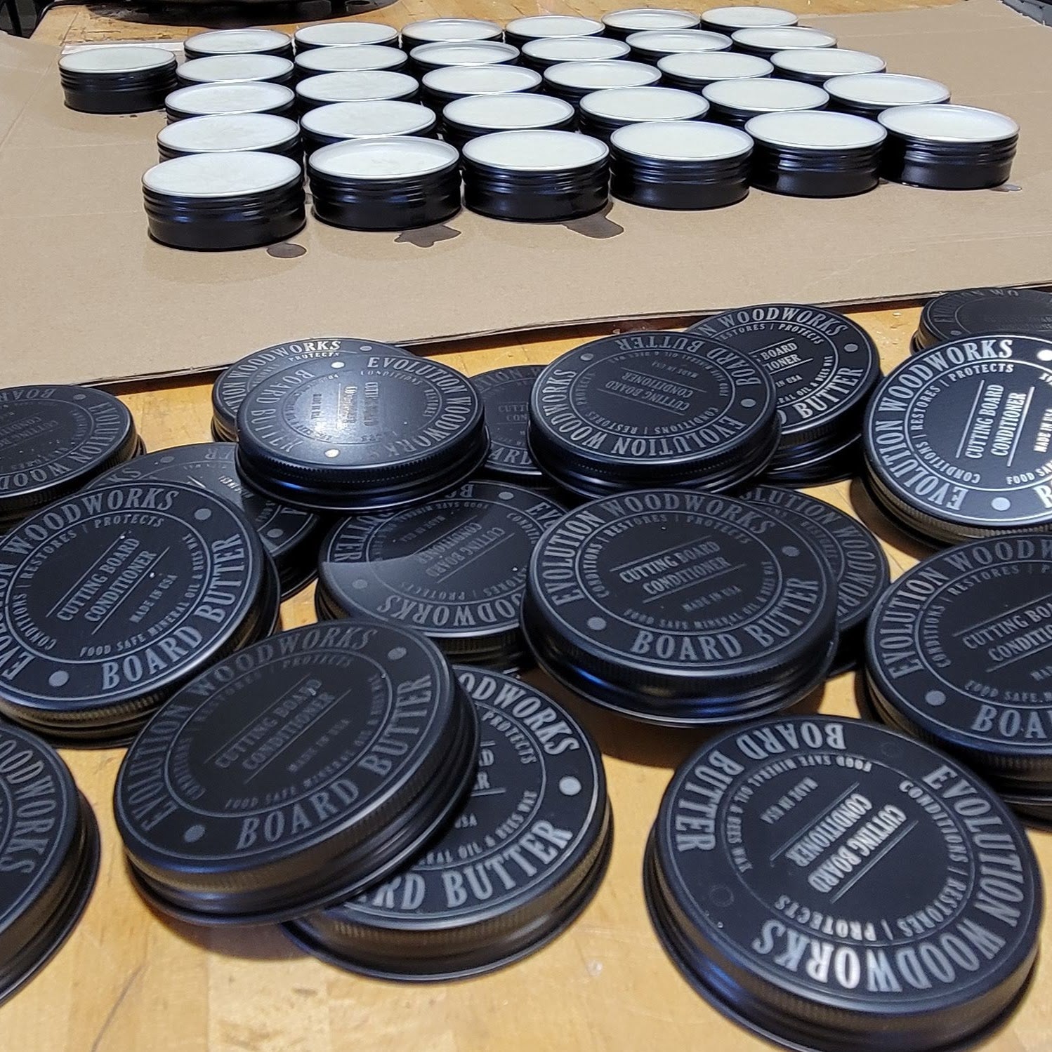 Stacks of lids in front of tins of fresh poured cutting board conditioner cooling in our workshop.